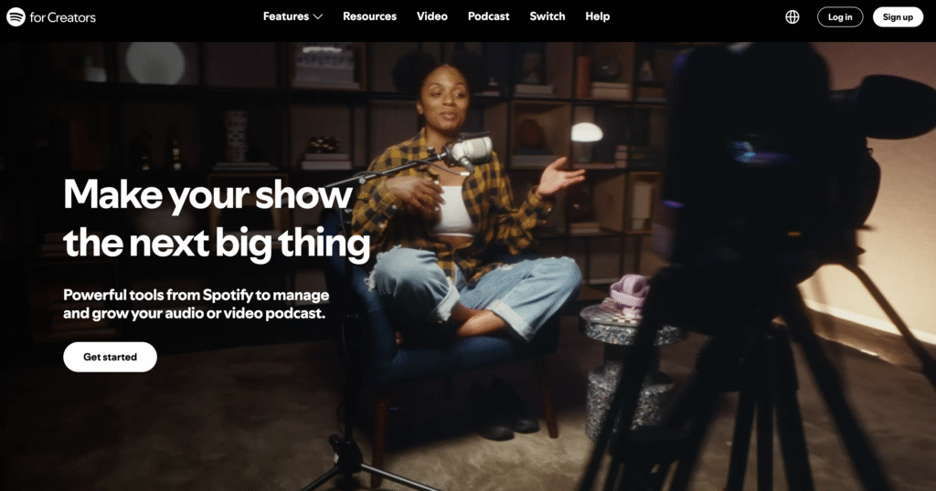 Uma pessoa está sentada em uma cadeira azul, gravando um podcast com um microfone, cercada por prateleiras e com uma câmera focada nela.